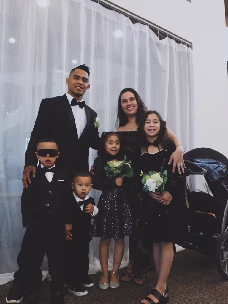 A formal family portrait featuring Kai Kamaka III in a black tuxedo and bowtie. He is standing with his wife and four children all dressed in formal black attire in front of a white draped background